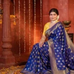 A woman wearing a royal blue Banarasi silk saree with golden motifs and traditional jewellery in a festive heritage-style setting
